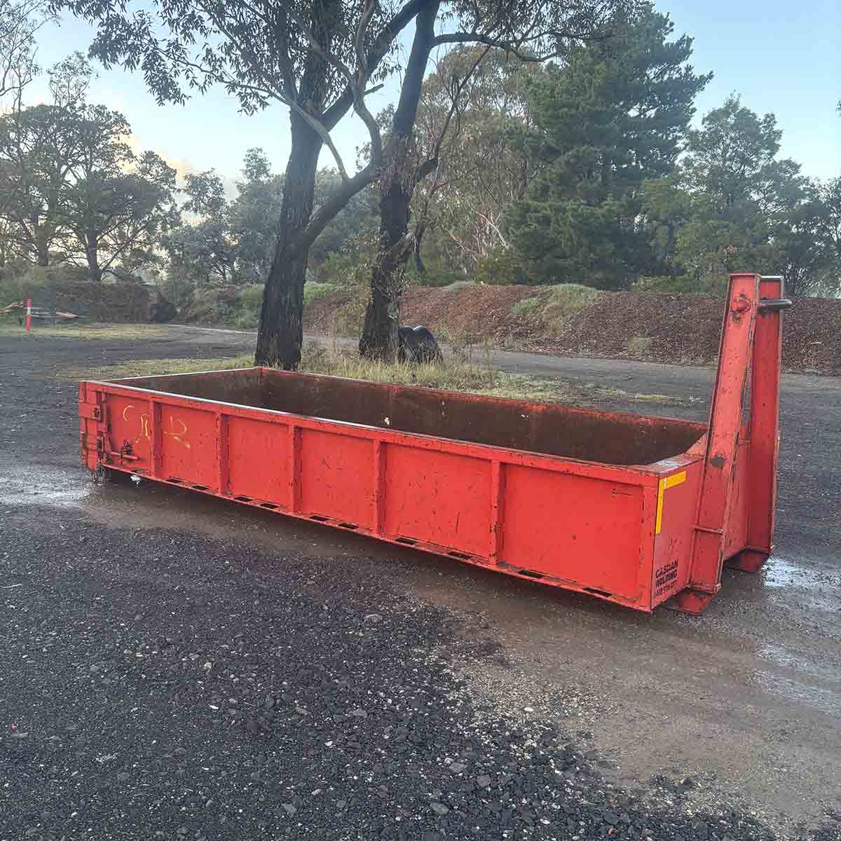 Red walk-in bin on a gravel yard under gum trees, square crop
